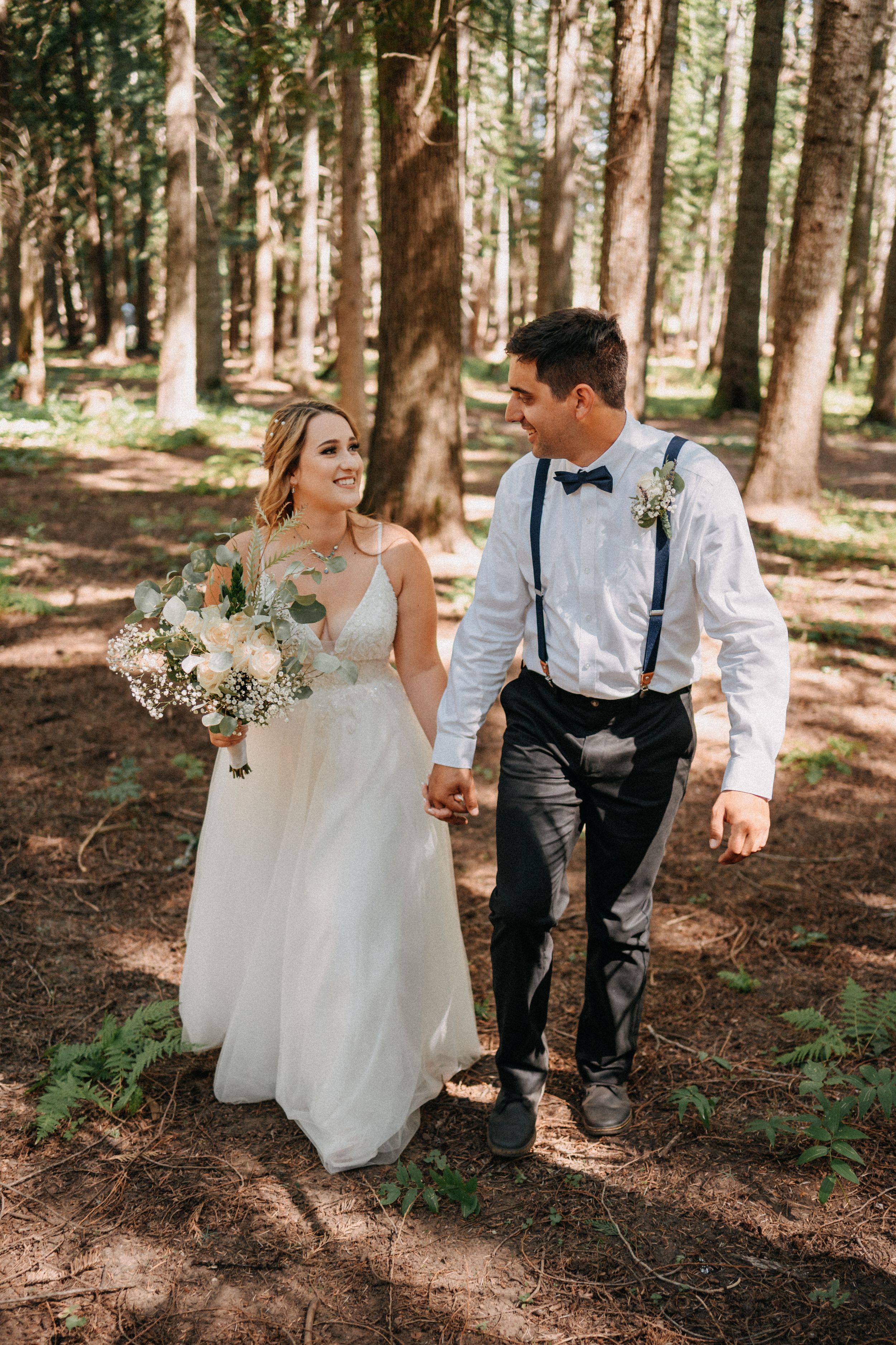 A couple walks hand in hand through a sunlit forest, wearing wedding attire with soft bouquet and rustic suspender style.