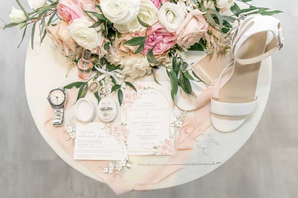 Wedding details flatlay with pink and white roses, wedding rings, invitations, and bridal shoes on blush surface.