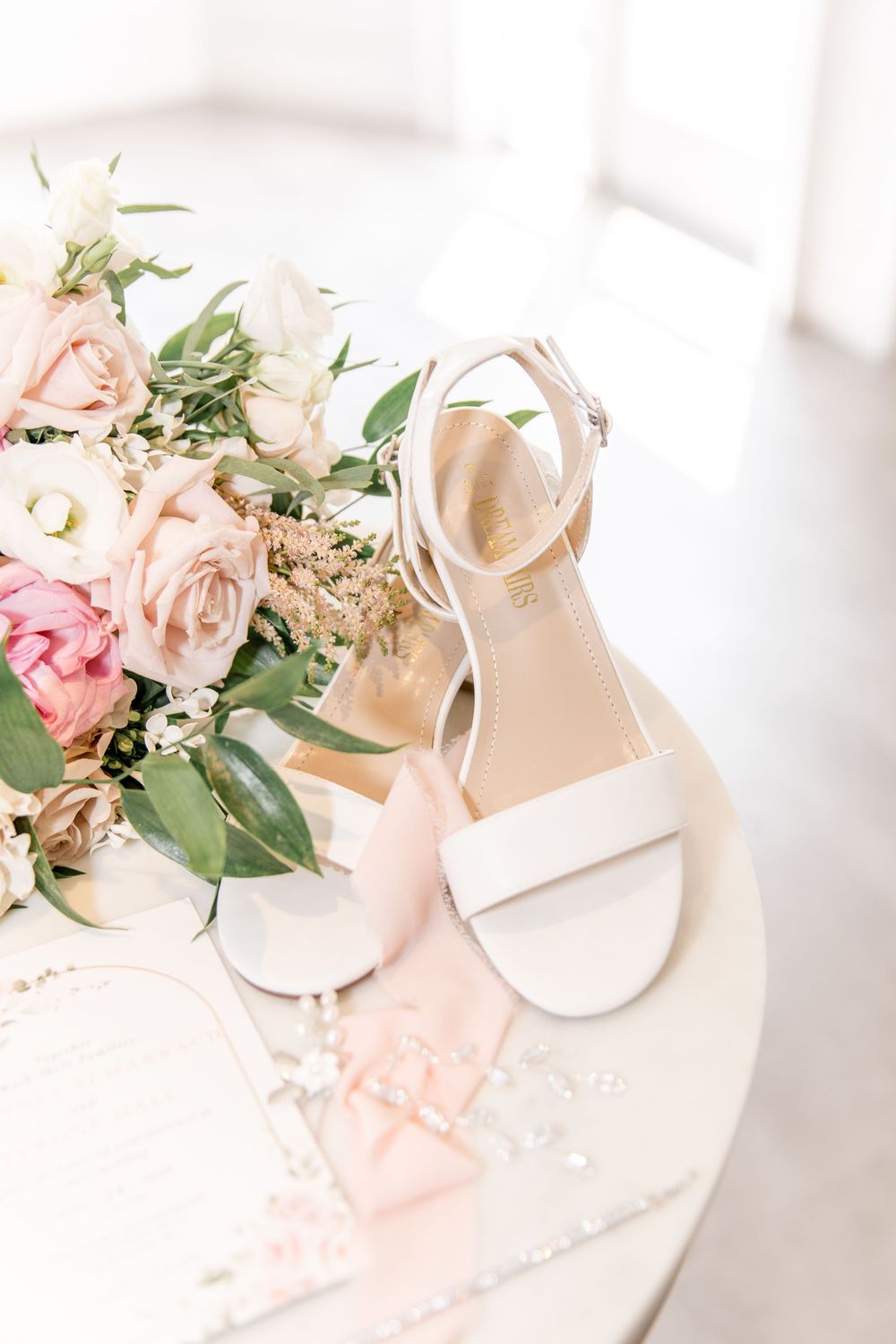 White and pink bridal accessories including shoes and delicate floral arrangements on a light background.
