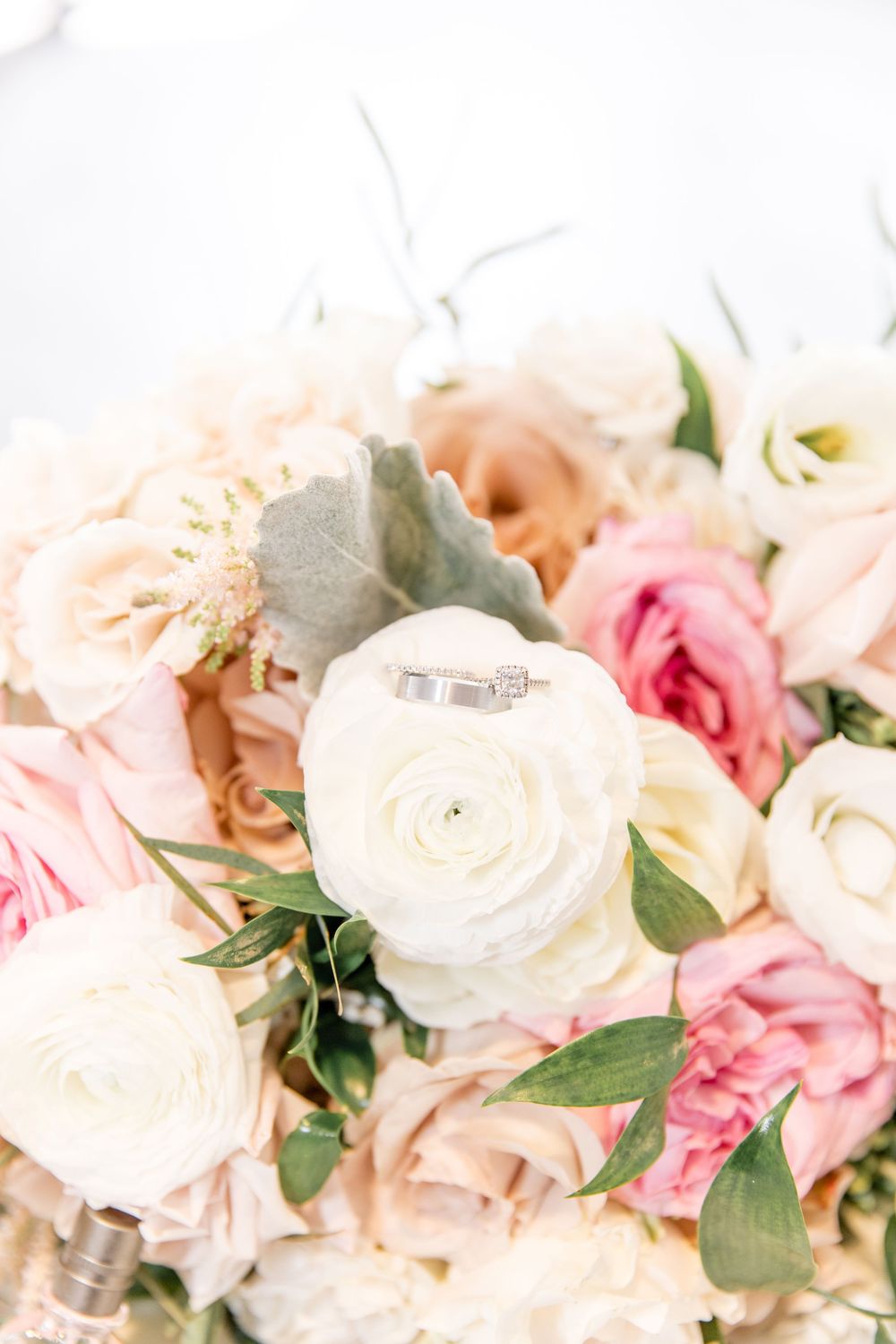 Wedding rings rest delicately on a white rose surrounded by pink and peach roses and greenery in a romantic bouquet.