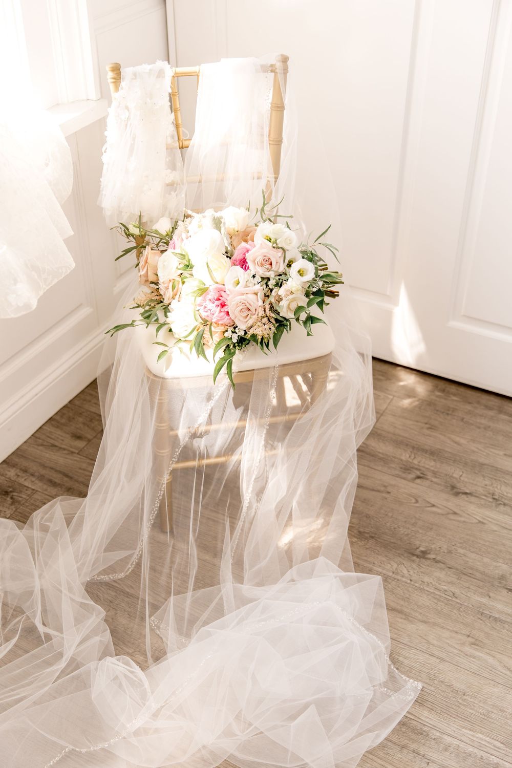 A delicate bridal bouquet of pink and white flowers rests on a chair draped with flowing white tulle veil.