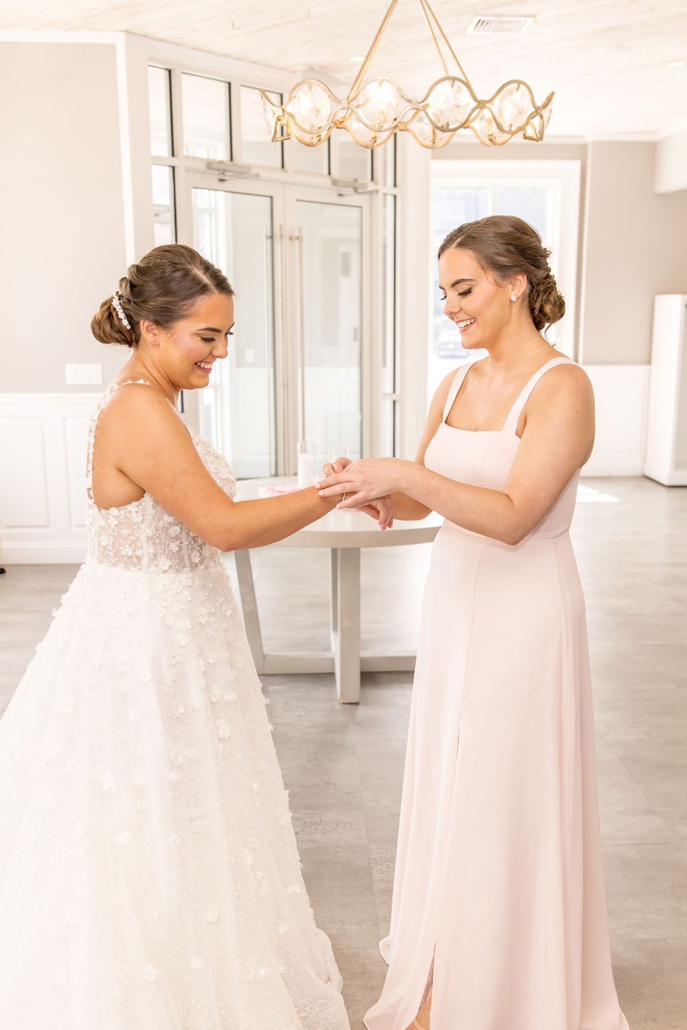 Two elegantly dressed attendees in formal gowns share a tender moment in a bright room with a chandelier.