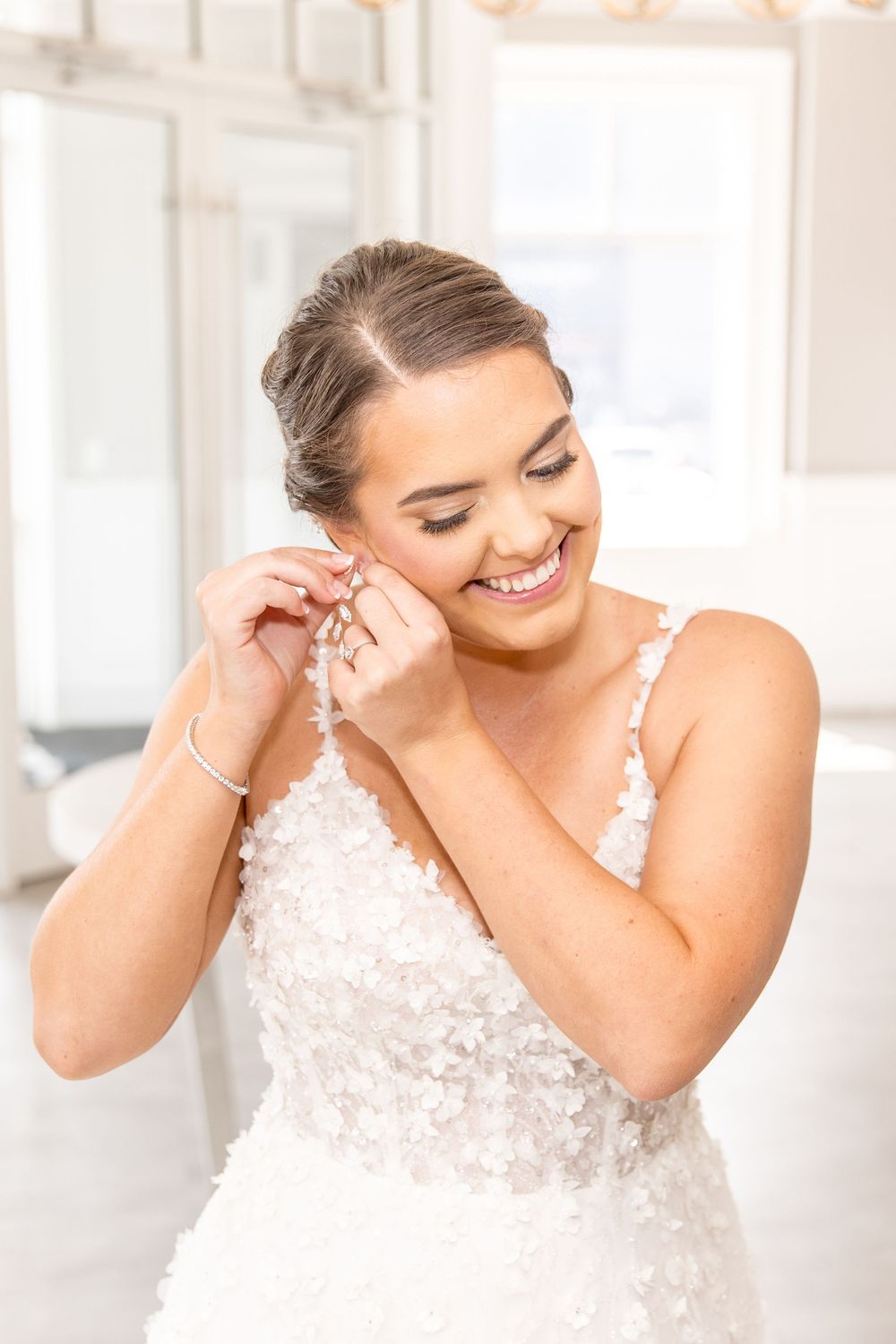 A bride in a lace wedding gown smiles while putting on earrings in a bright sunlit room.