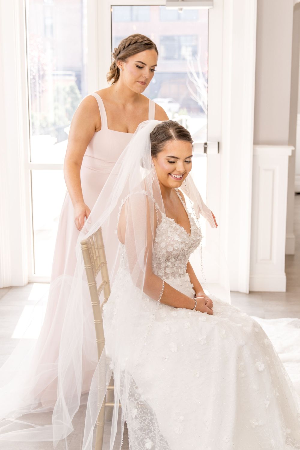 Two attendees in matching formal gowns share an intimate moment during wedding preparations in a sunlit room.