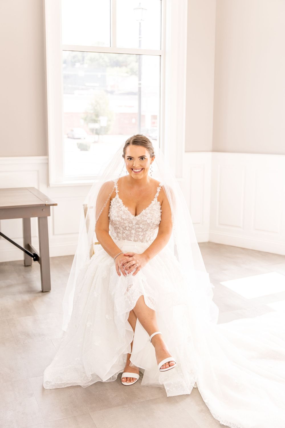 A bride in a beautiful lace gown and white sandals sits gracefully by a bright window in a light-filled room.