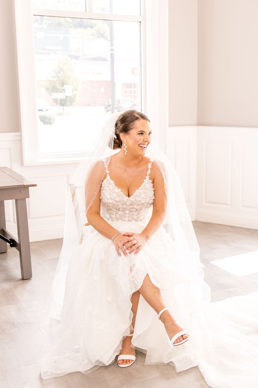 A bride in a white lace wedding dress and sandals sits gracefully in a bright sunlit room.