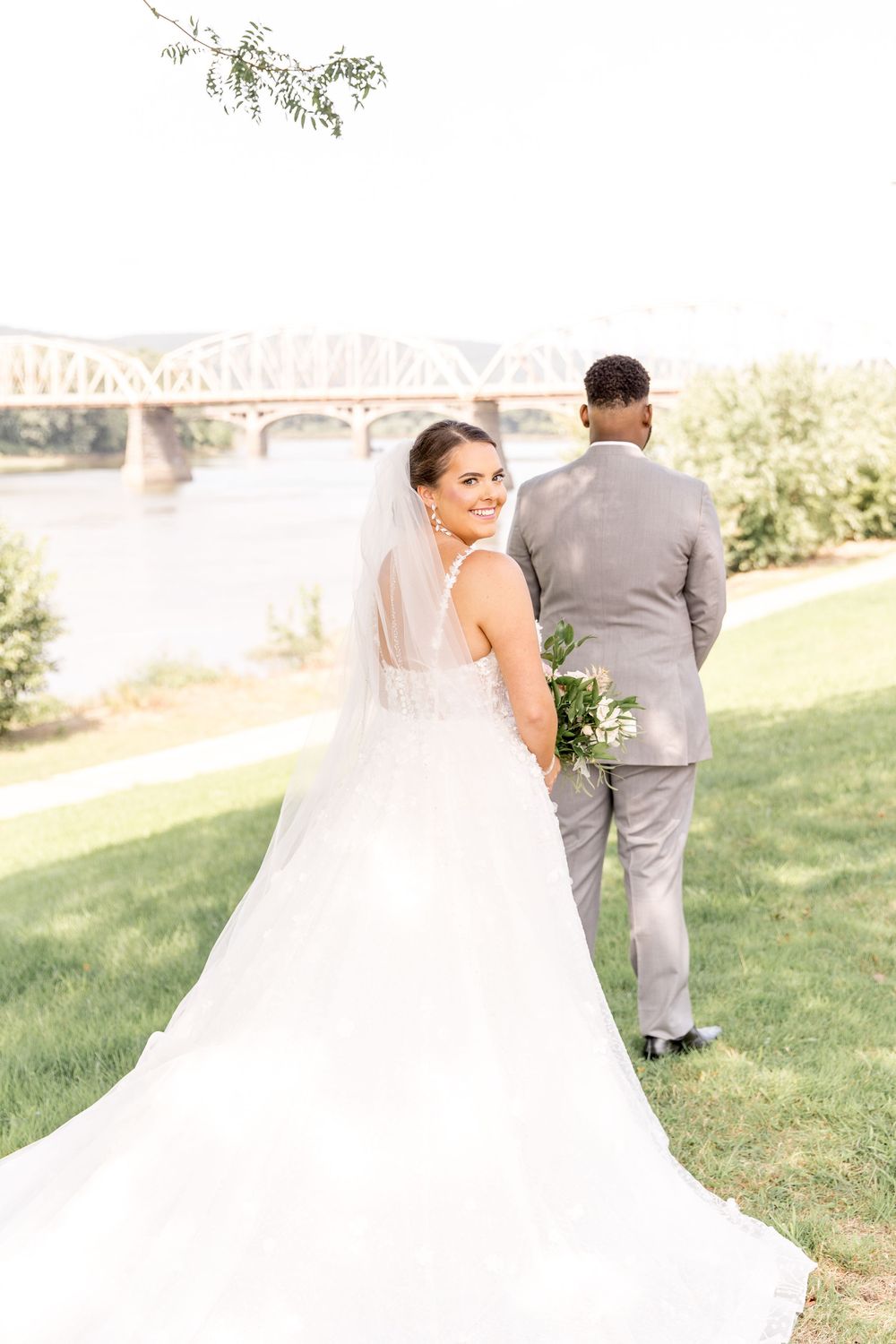 Bride in flowing white dress looks back at camera while standing with partner near bridge on grassy hill during wedding.