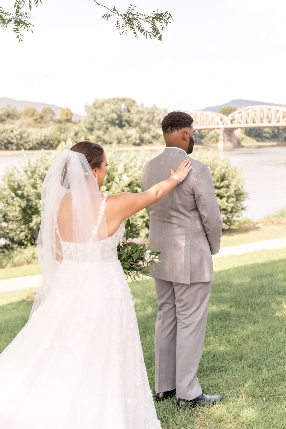 A bride in a white wedding dress approaches their partner wearing a gray suit on a grassy hill overlooking a bridge.