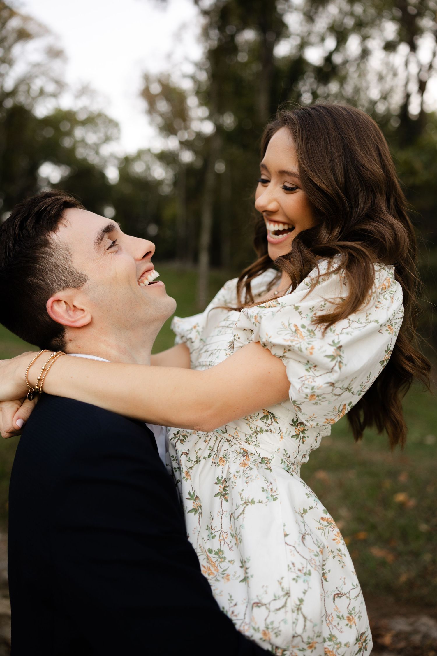 A couple shares a joyful moment outdoors as they embrace and laugh together against a natural wooded backdrop.