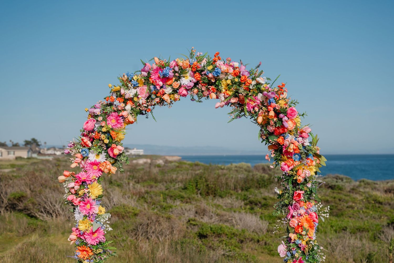 Wedding photo showing a bride in a lace dress beside a colorful floral arch with pink and yellow blooms against glass windows.