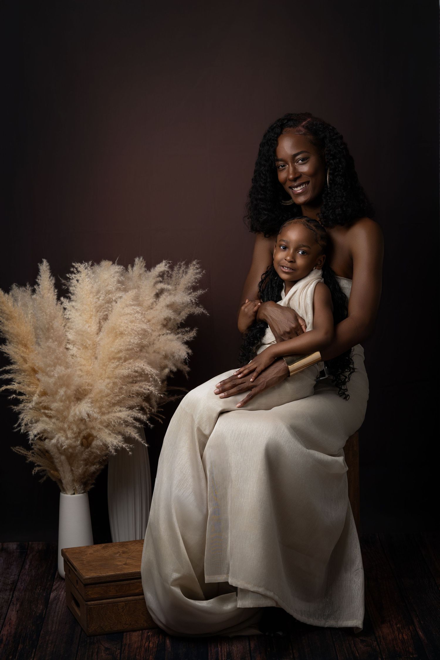 An intimate portrait of two people in white dresses sitting together against a dark backdrop with pampas grass decor.