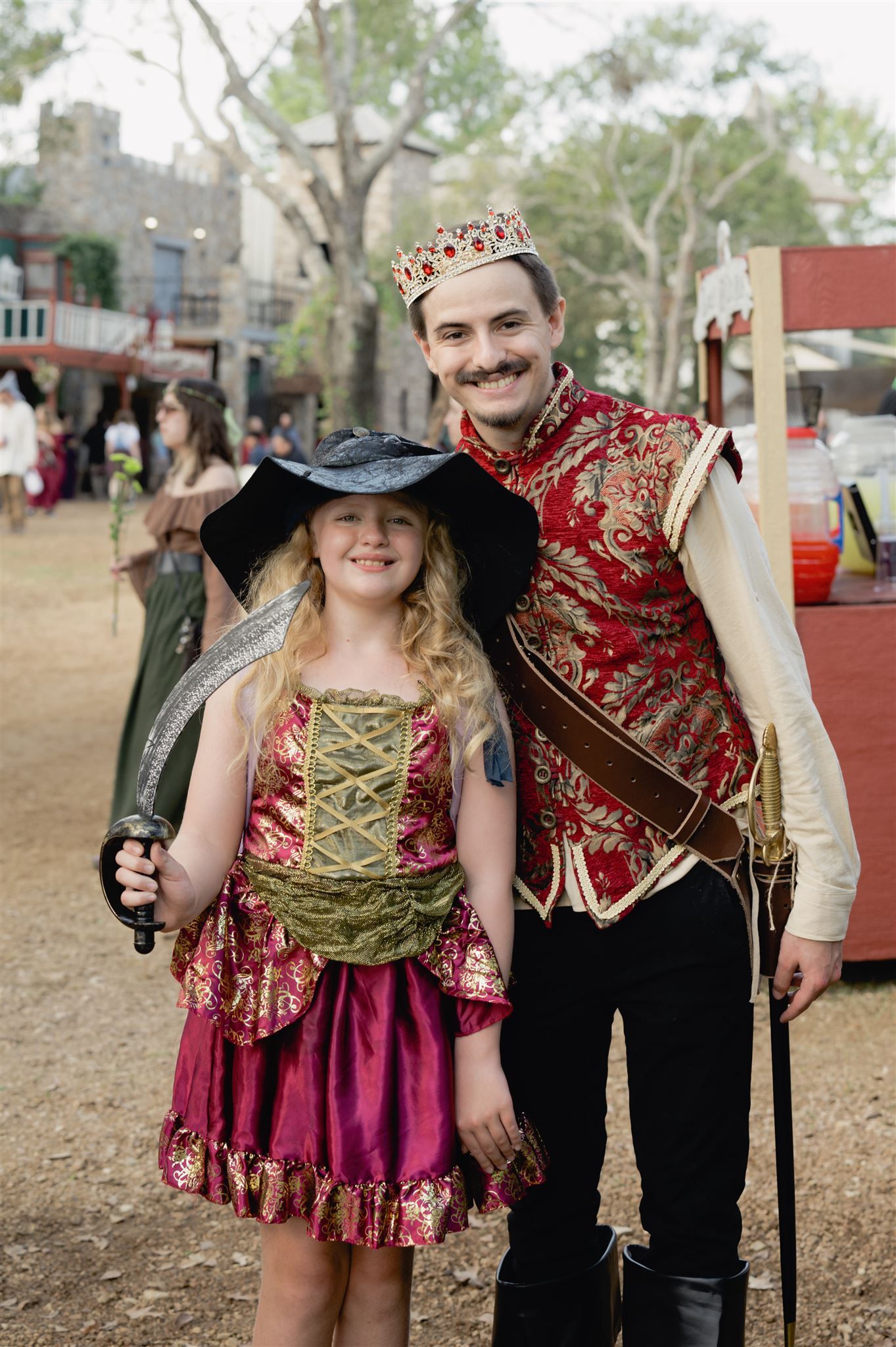 Two people in Renaissance festival costumes pose together, wearing fancy period dress and accessories.