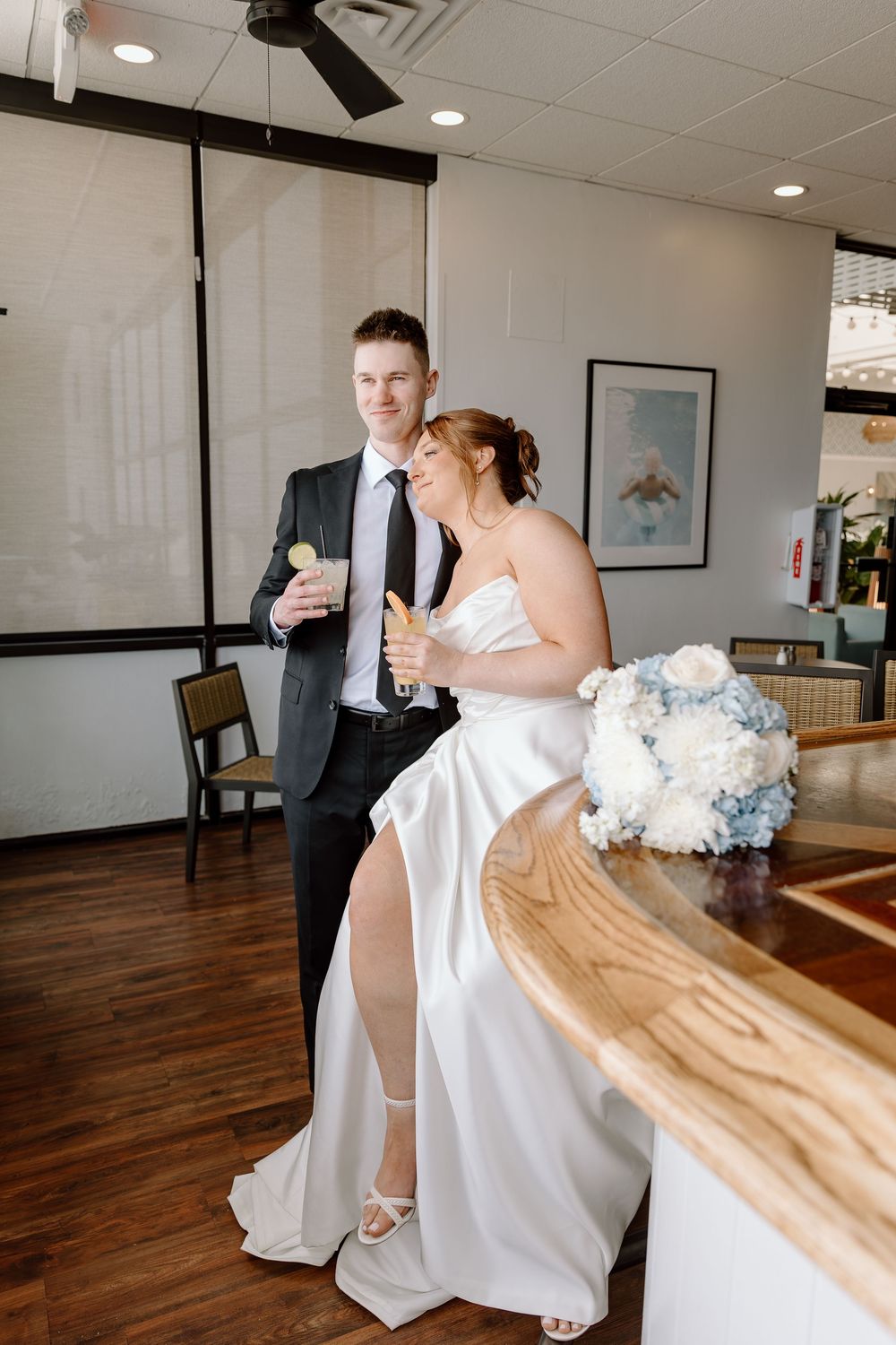 The bride in a white wedding dress sits at a wooden table while sharing a sweet laughter-filled moment with guests.
