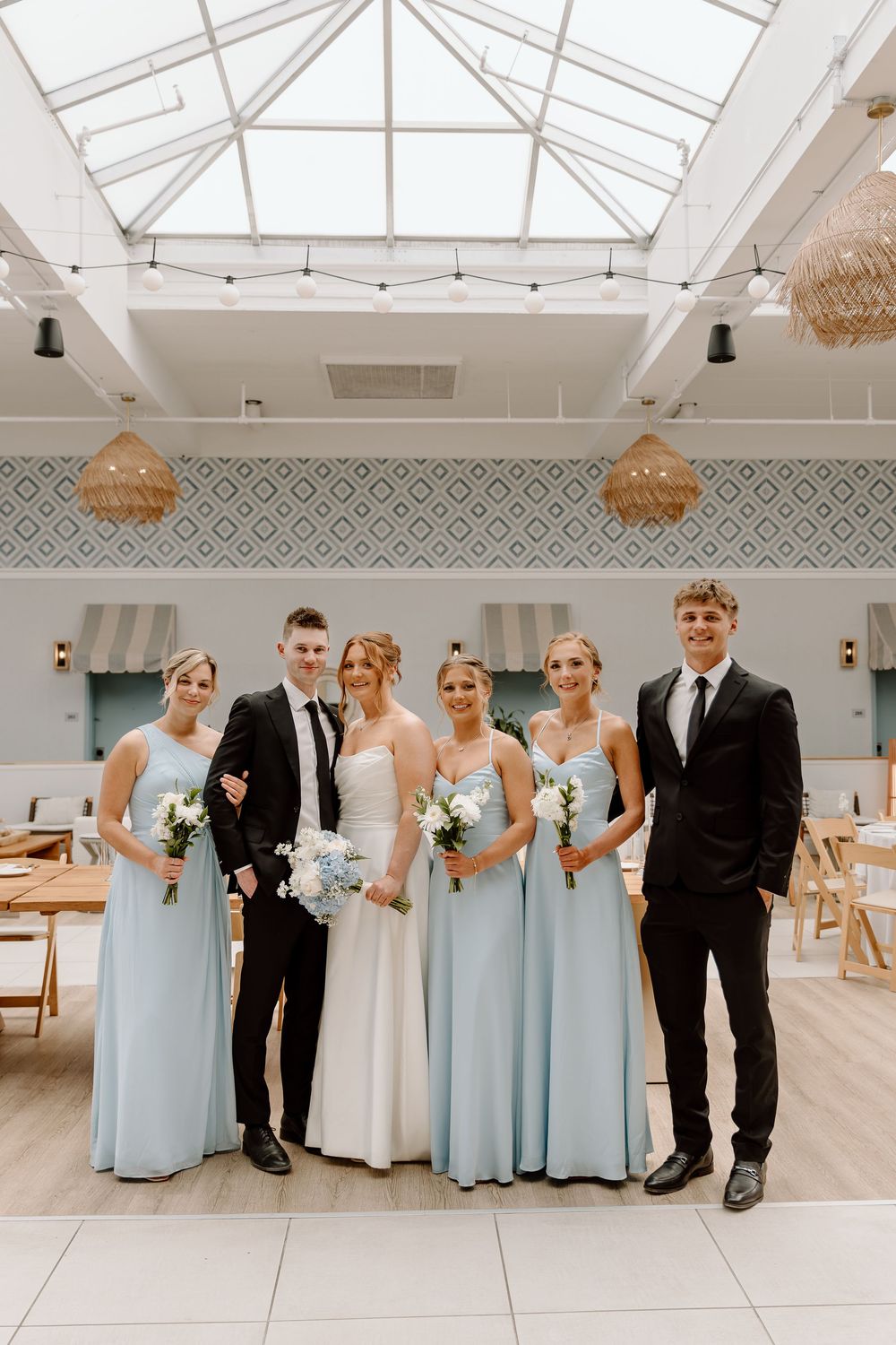 Wedding party in pale blue and black attire poses in modern white venue with skylights and pendant lights.