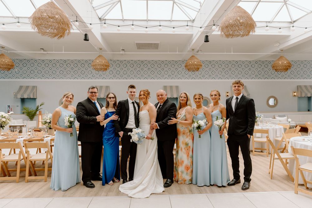 A wedding party in light blue bridesmaid dresses and formal attire poses for group photos in a bright indoor venue.