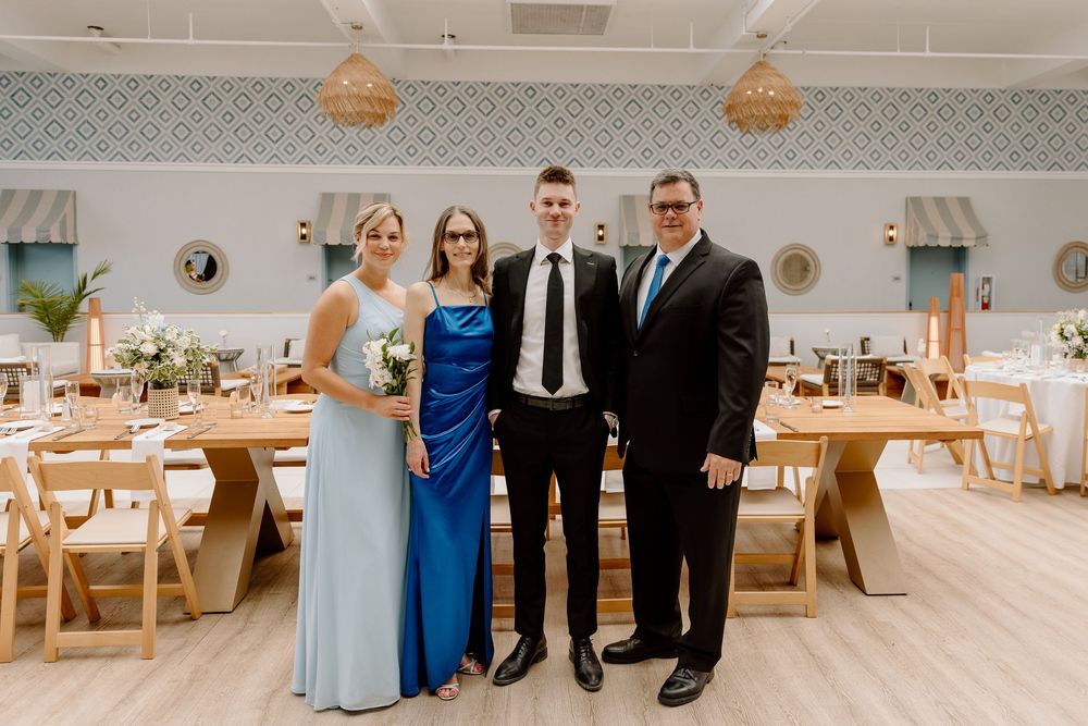 Four people in formal attire pose elegantly in a rustic wedding reception venue with wooden tables and decorative lighting.