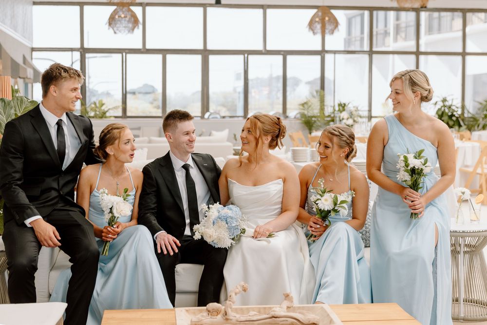 Wedding party shares joyful laughter while seated in a row at a bright reception venue decorated with floral arrangements.