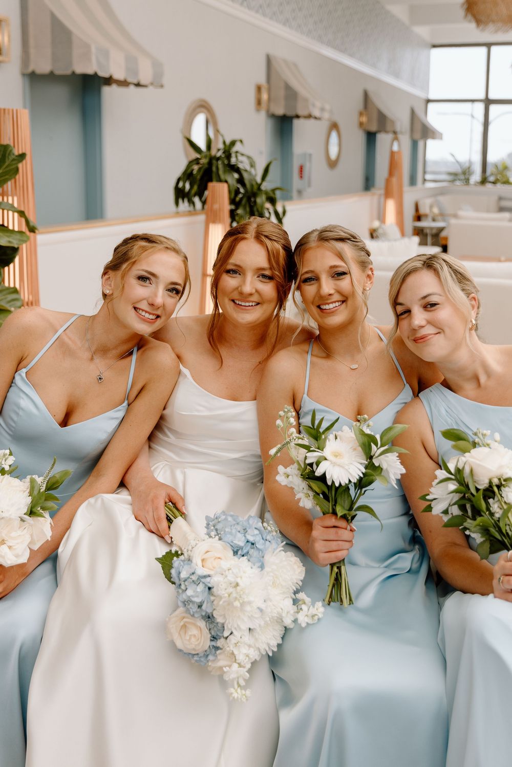 A bridal party wearing white and light blue silk dresses hold delicate bouquets of white and blue flowers at a wedding.