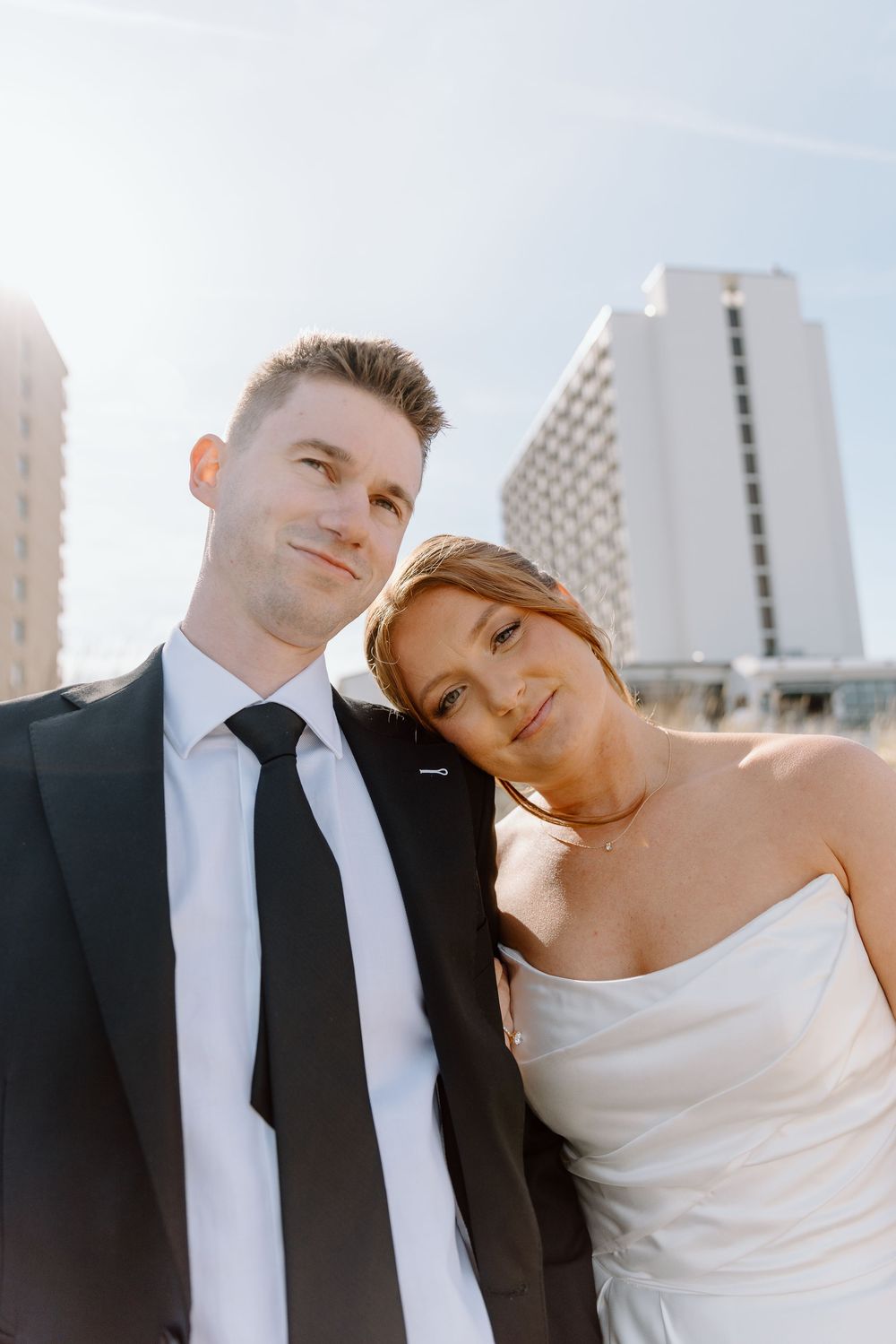 A couple in wedding attire pose together outdoors with a tall modern hotel building visible in the background.