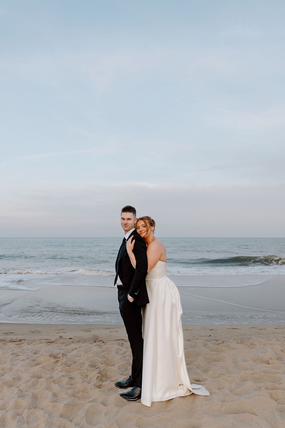 A couple shares an intimate moment on a serene beach with the ocean in the background and cloudy skies above.