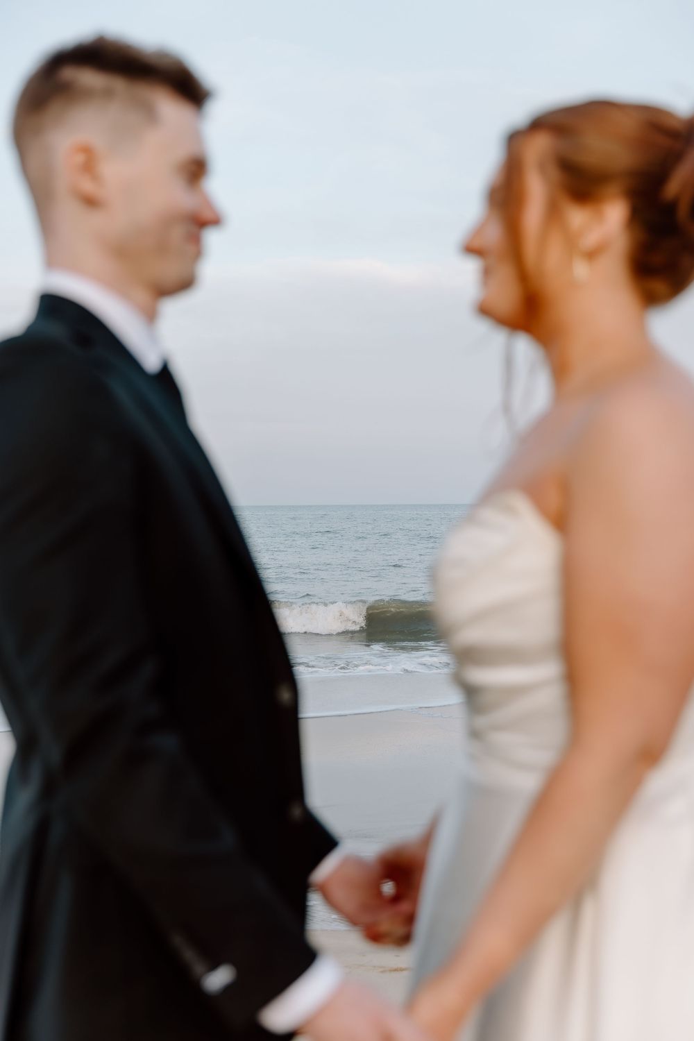 A romantic beach wedding scene with a couple in formal attire standing by the ocean at sunset.
