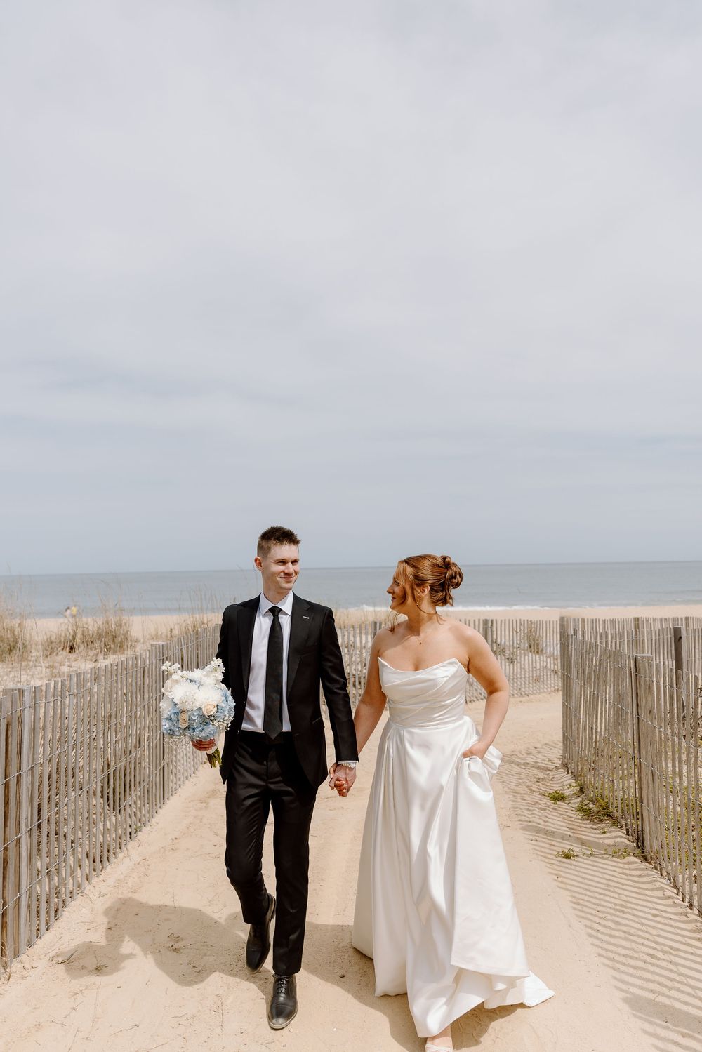 A couple in wedding attire holding hands while walking on a sandy beach path with ocean and dunes in background.