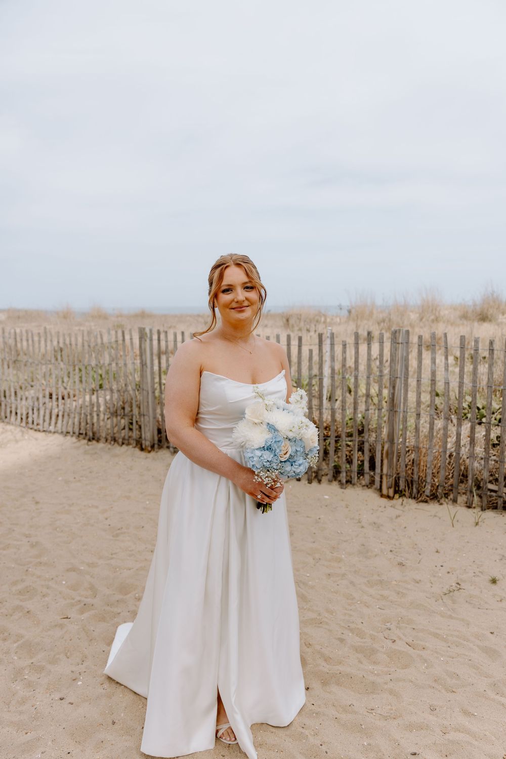 A bride in a flowing white gown holds a light blue bouquet while standing on a sandy beach with a wooden fence behind her.