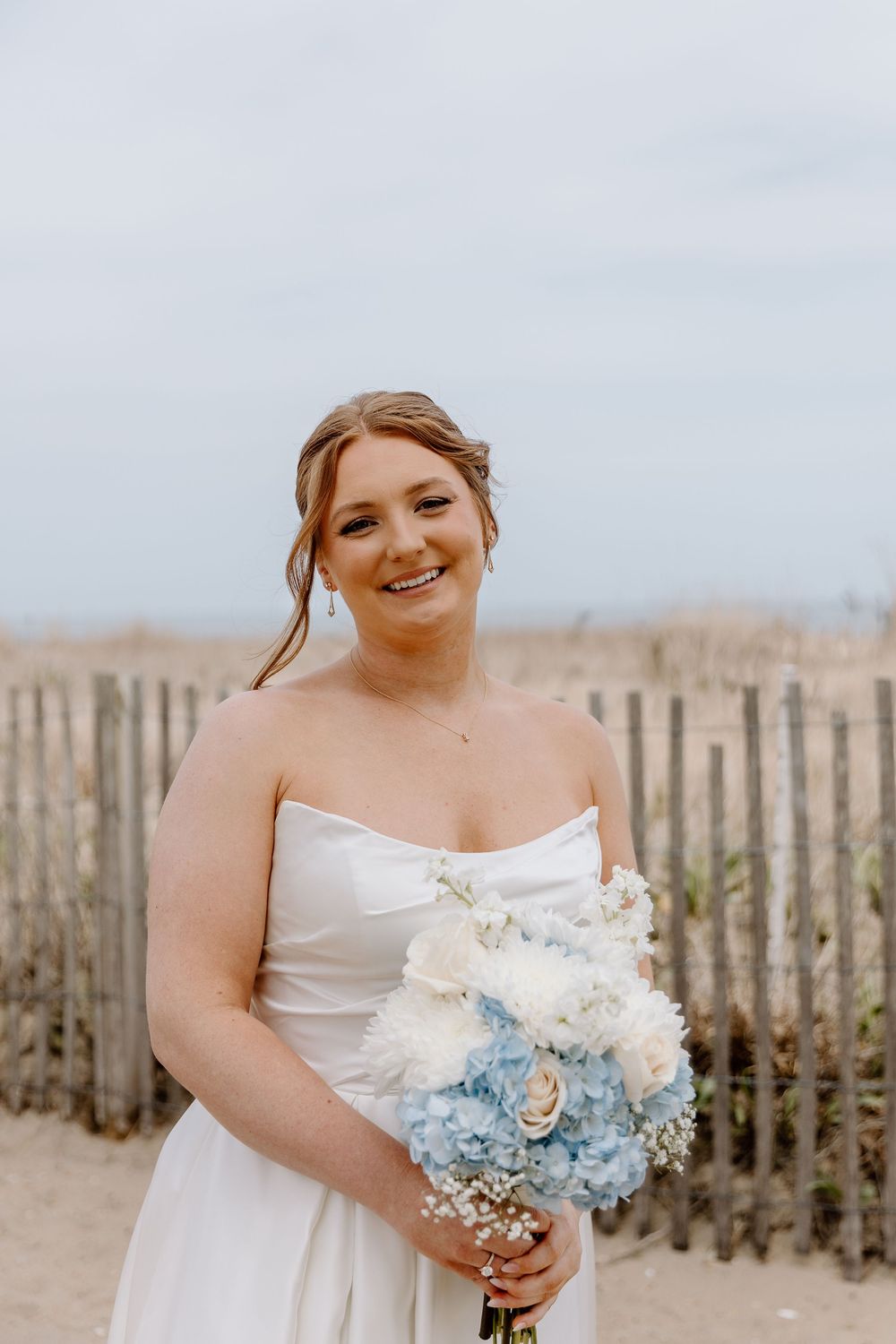 A smiling bride in a white strapless gown holds a blue and white bouquet on a beach with a wooden fence behind her.