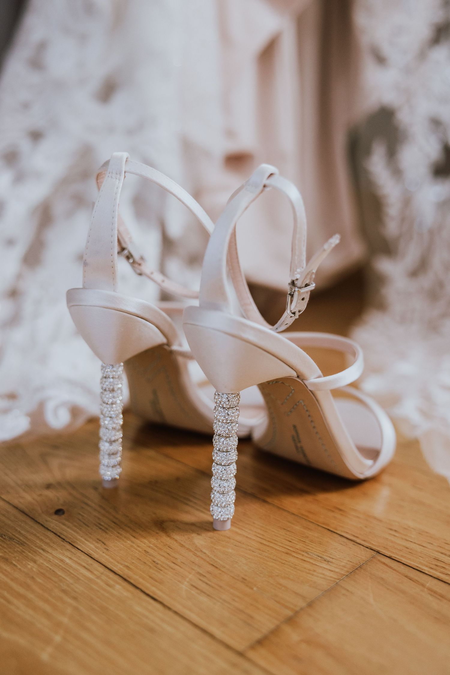 Elegant white bridal heels with crystal-embellished heels displayed on a wooden floor showcasing their sparkly details.