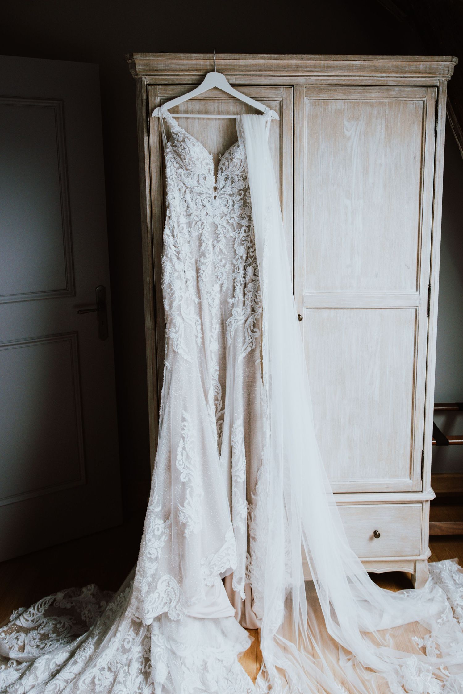A delicate lace wedding dress with intricate beading and long train hangs from a white hanger against an antique wardrobe.