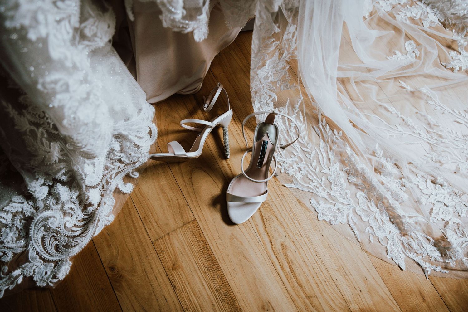Elegant silver wedding heels next to flowing white lace dress on wooden floor.