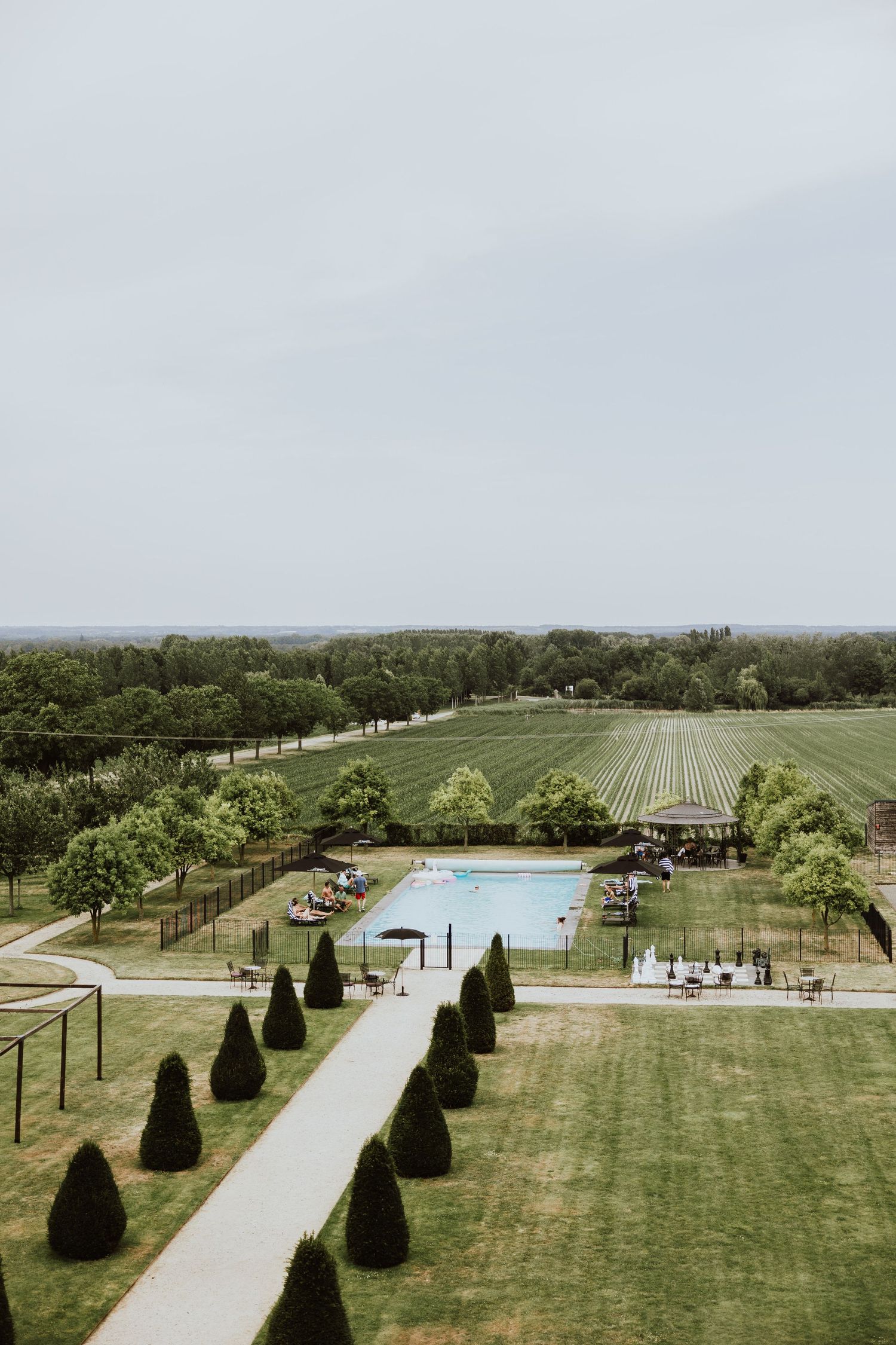 Aerial view of a manicured estate garden with swimming pool, lined cypress trees, and agricultural fields in background.