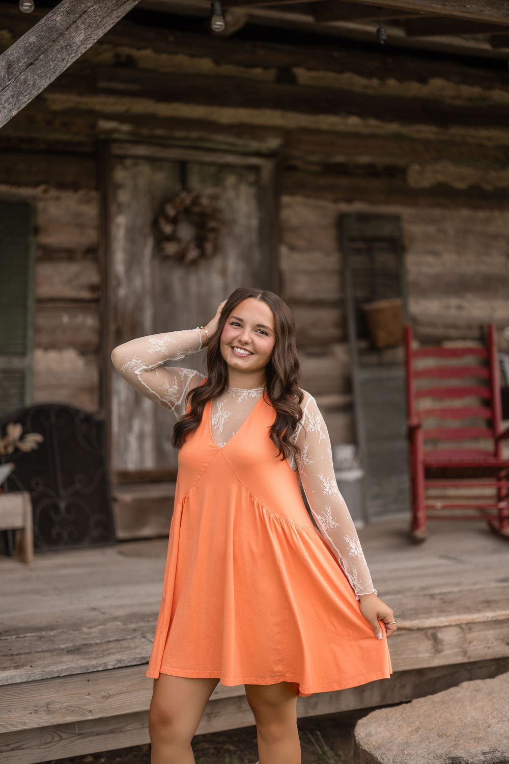 A woman in an orange sundress and lace cardigan poses on a rustic wooden porch of a log cabin.