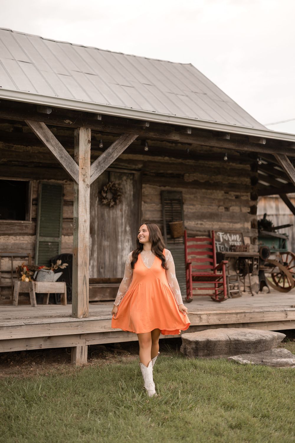 Woman in flowy orange dress and white boots poses in front of rustic wooden cabin porch with vintage decor.