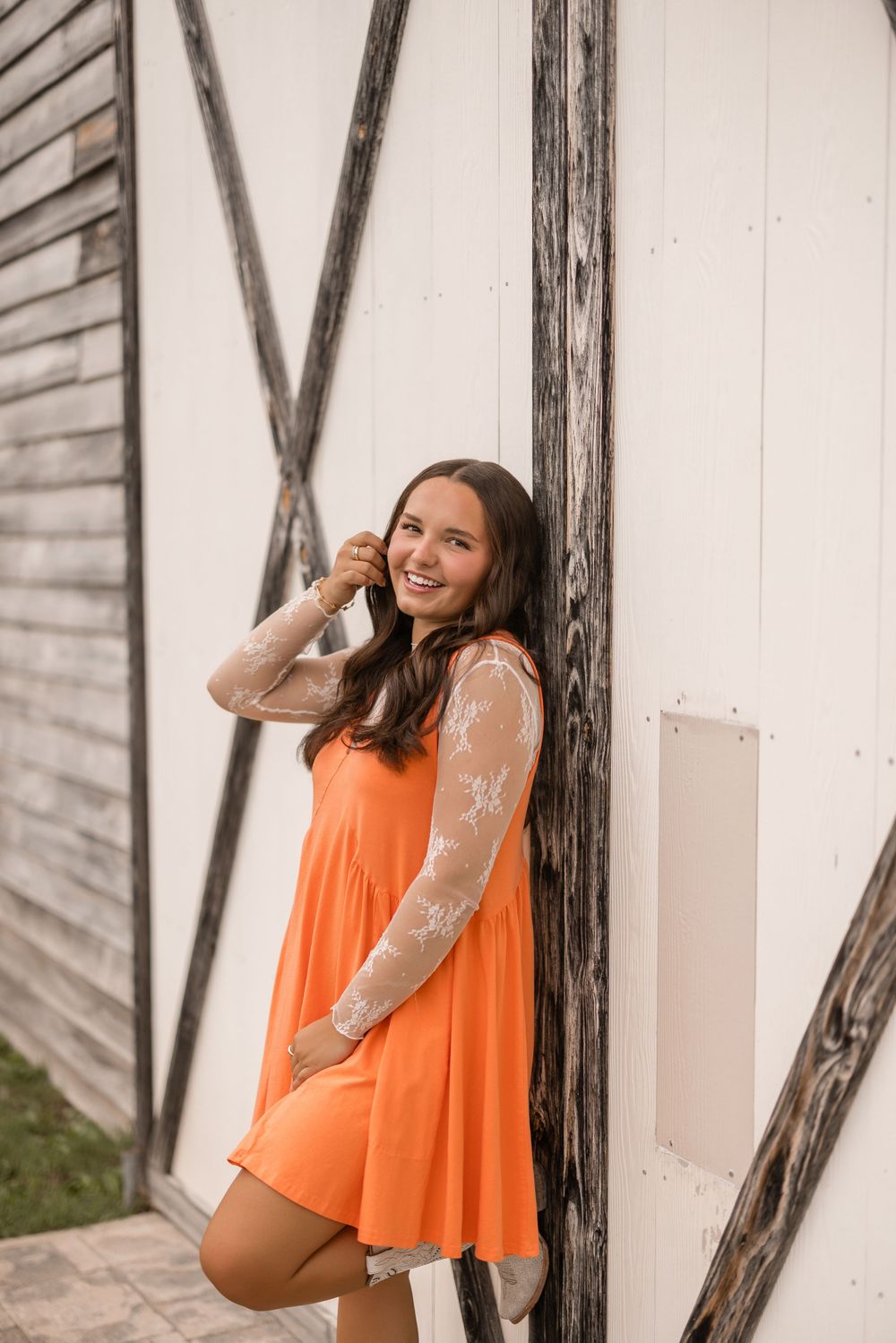 A person in an orange dress and white lace sleeves poses against a rustic wooden barn wall.