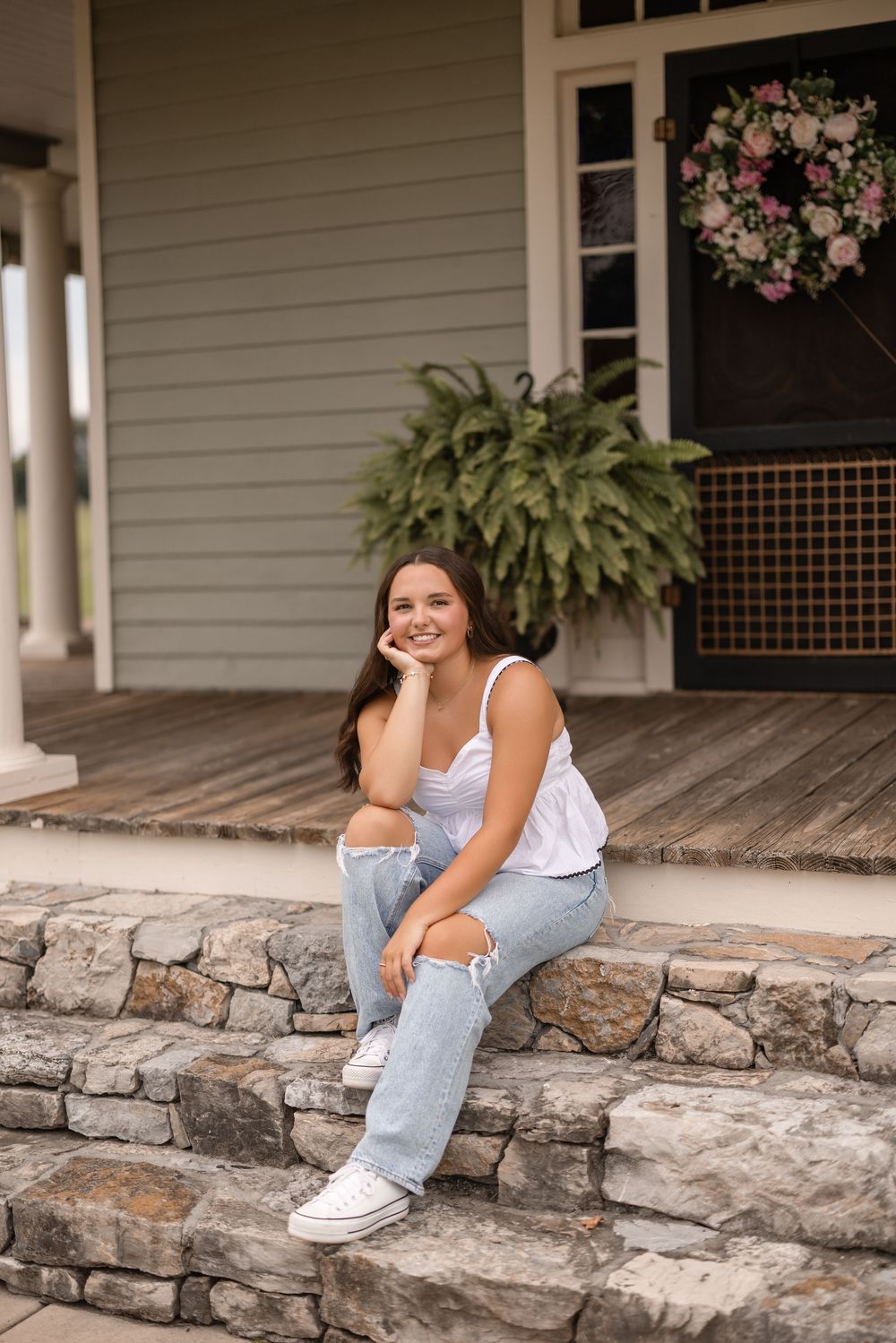 A person in white sneakers and jeans sits casually on rustic stone steps of a front porch with potted plants.