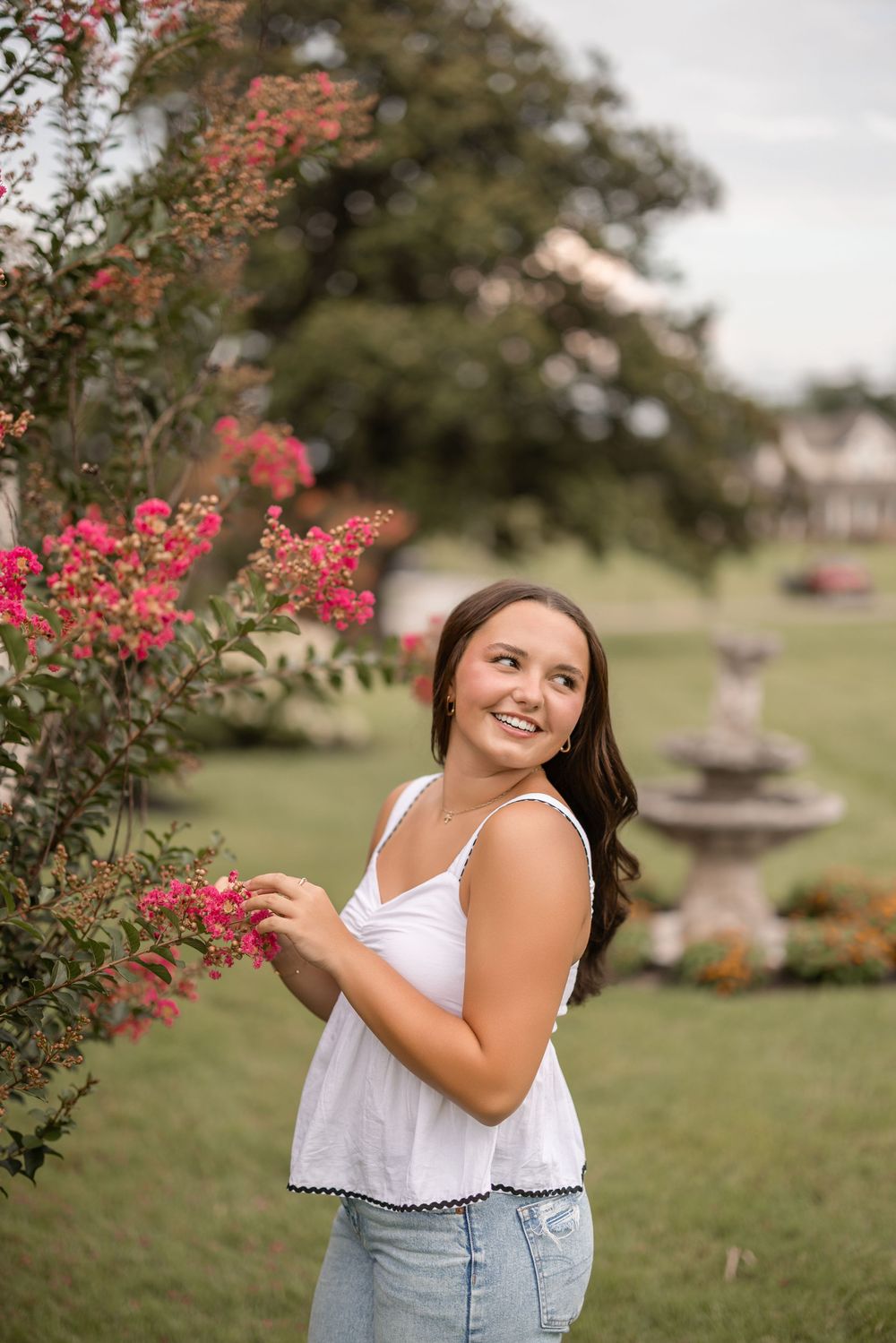 A figure in a white top and denim skirt poses near blooming pink Crape Myrtle flowers on a sunny summer day.