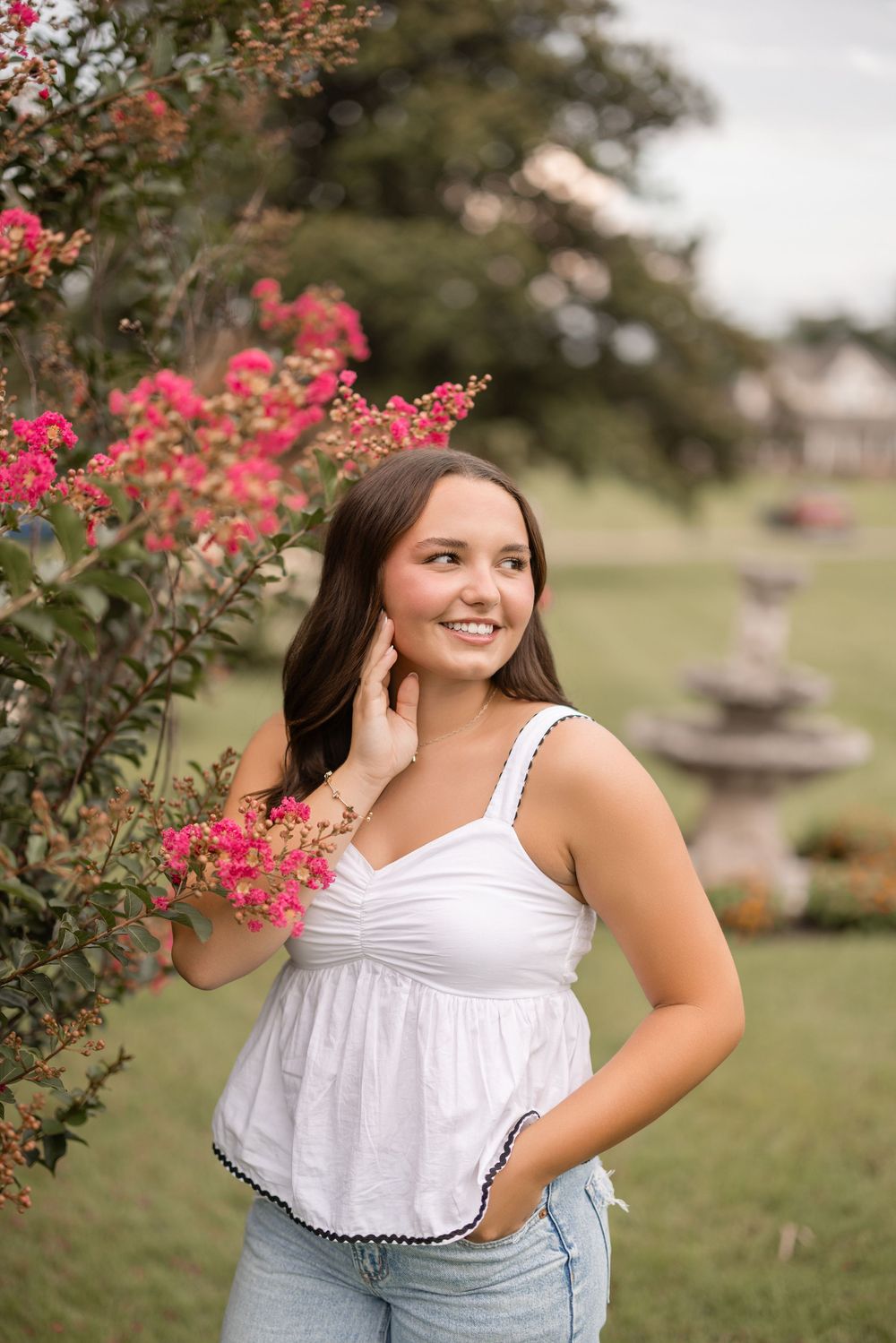 Pink flowering shrubs frame a sunny garden scene with someone posing in a white top against a natural backdrop.
