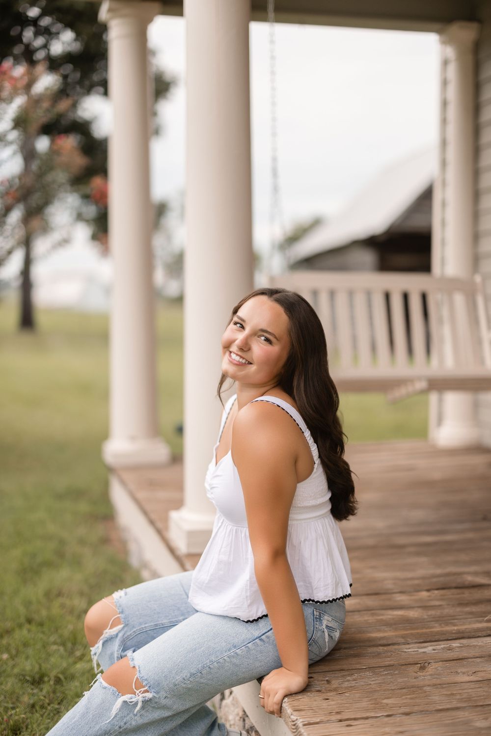A woman in ripped jeans and white top sits casually on a wooden porch with white columns on a sunny day.