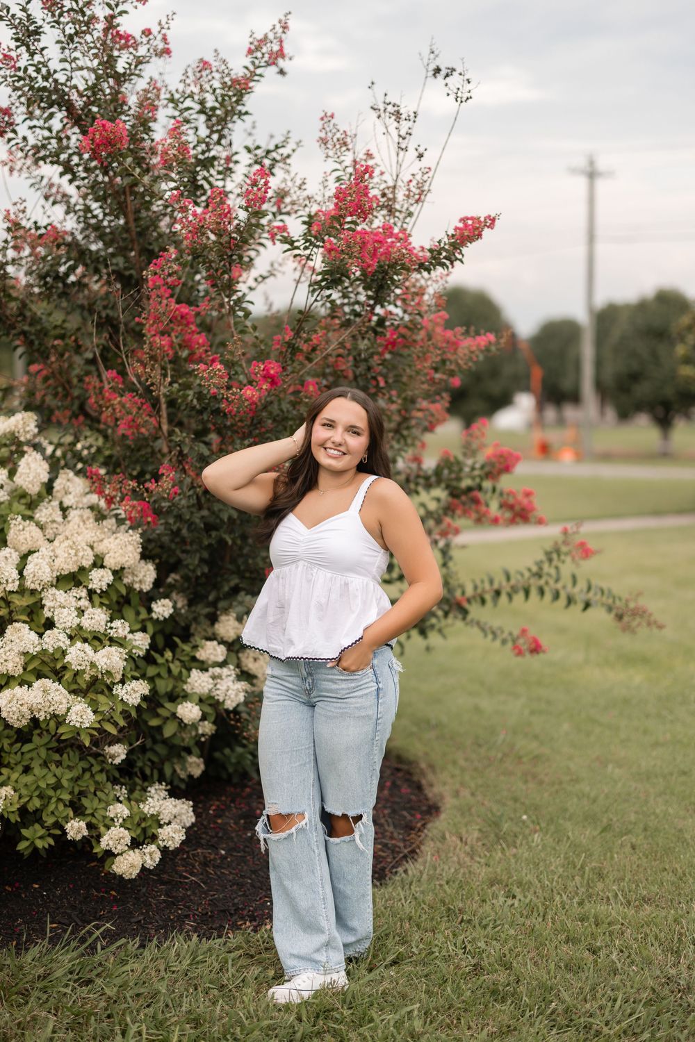 A figure poses outdoors in a white top and ripped jeans near flowering crepe myrtle and hydrangea bushes.