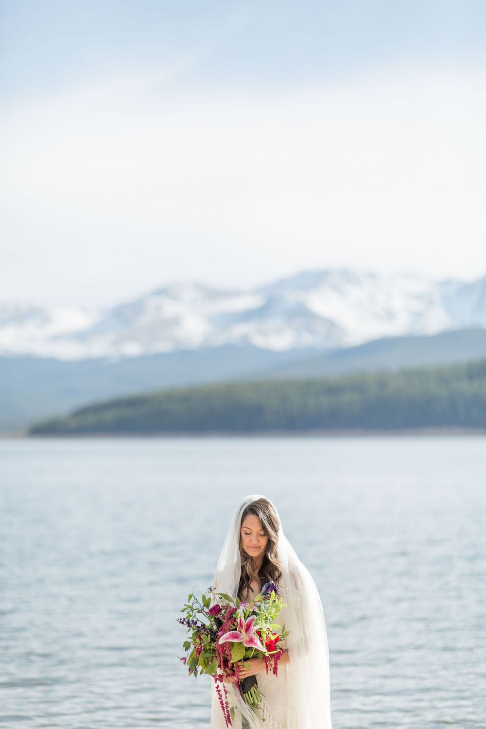 A person in a flowing white dress holds a vibrant bouquet against a mountainous lake backdrop.