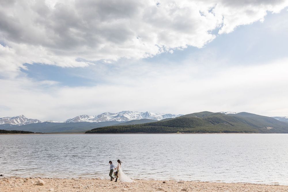 A serene mountain lake landscape with a distant snow-capped peaks and two figures walking along the sandy shoreline.