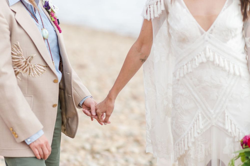 Couple in light-colored wedding attire holds hands while standing on sandy beach during sunset ceremony.