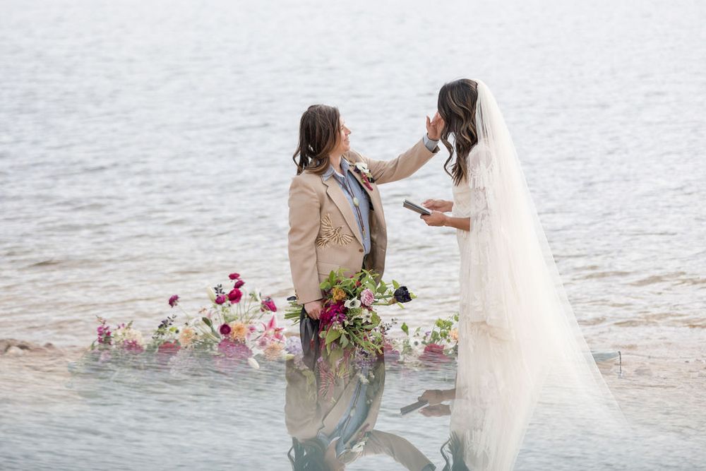 Two figures stand in a lake holding flowers wearing formal attire during a romantic photoshoot at sunset.