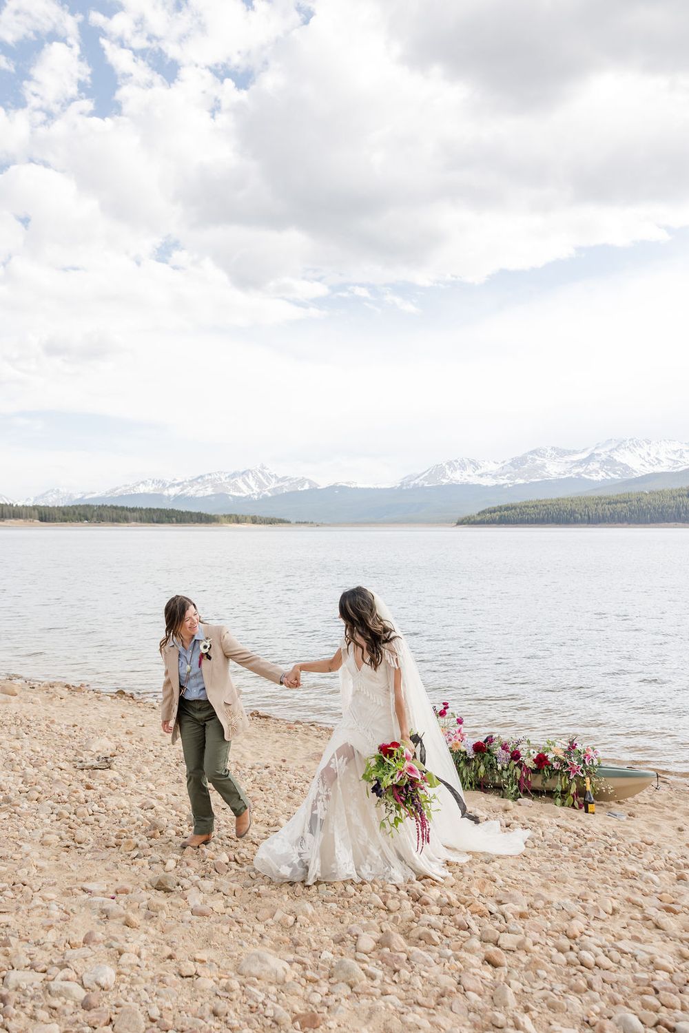 Two people in wedding attire hold hands while standing on a rocky beach shore with mountains and lake in background.