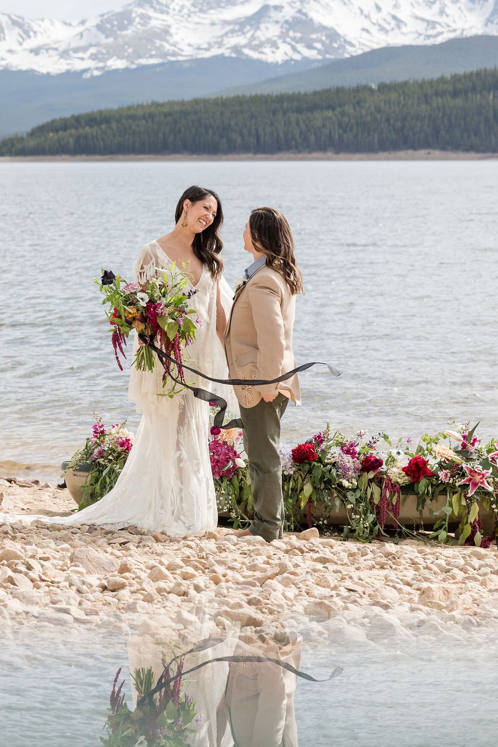 A wedding ceremony takes place on a rocky beach shore with snow-capped mountains and a lake in the background.
