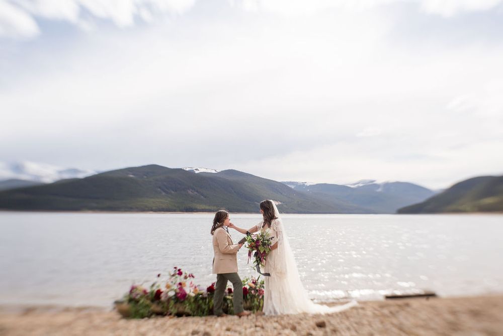 A wedding ceremony takes place on the shore of a scenic mountain lake with mountains rising in the background.