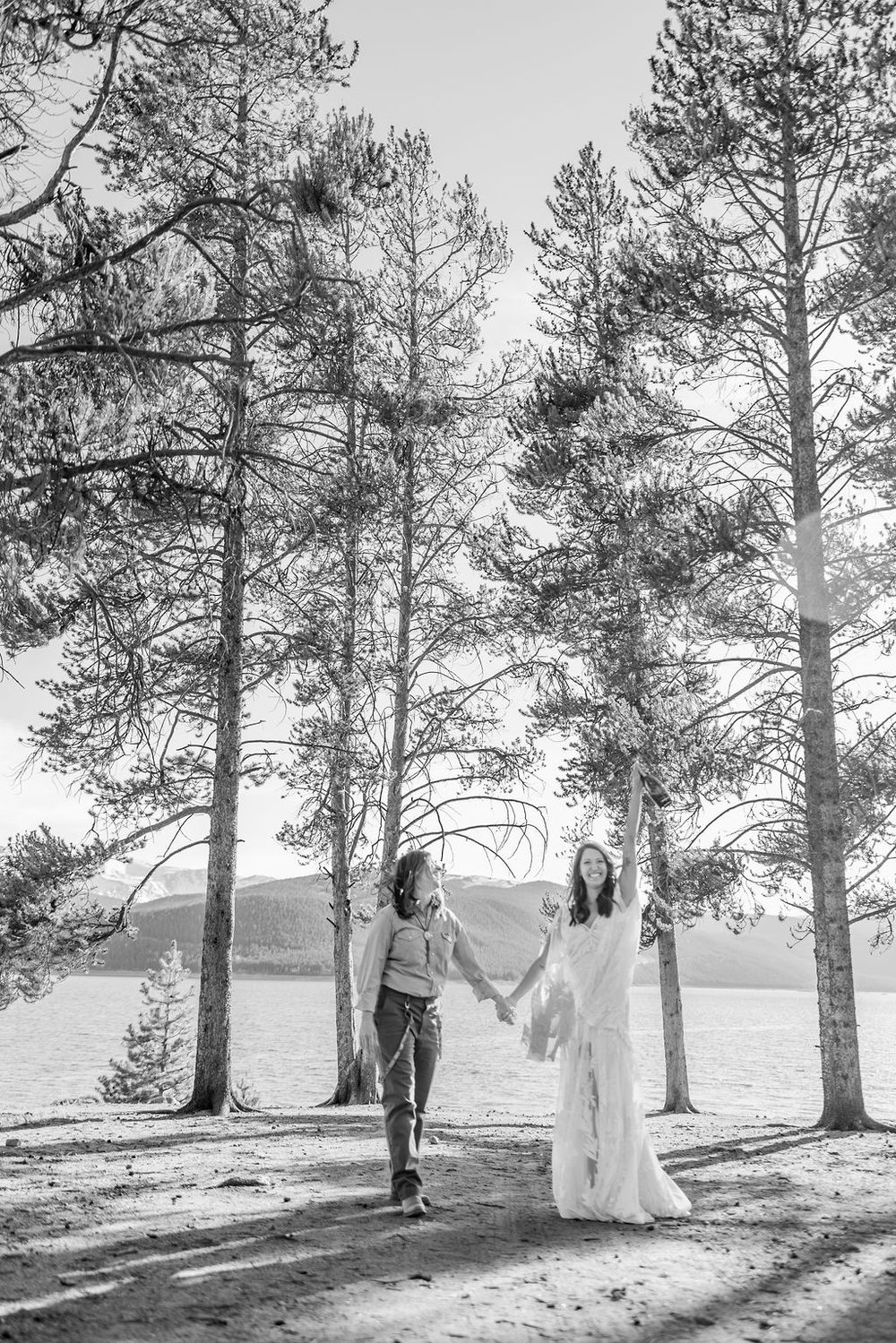 A romantic black and white photo of a couple holding hands among tall pine trees by a lake on a sunny day.