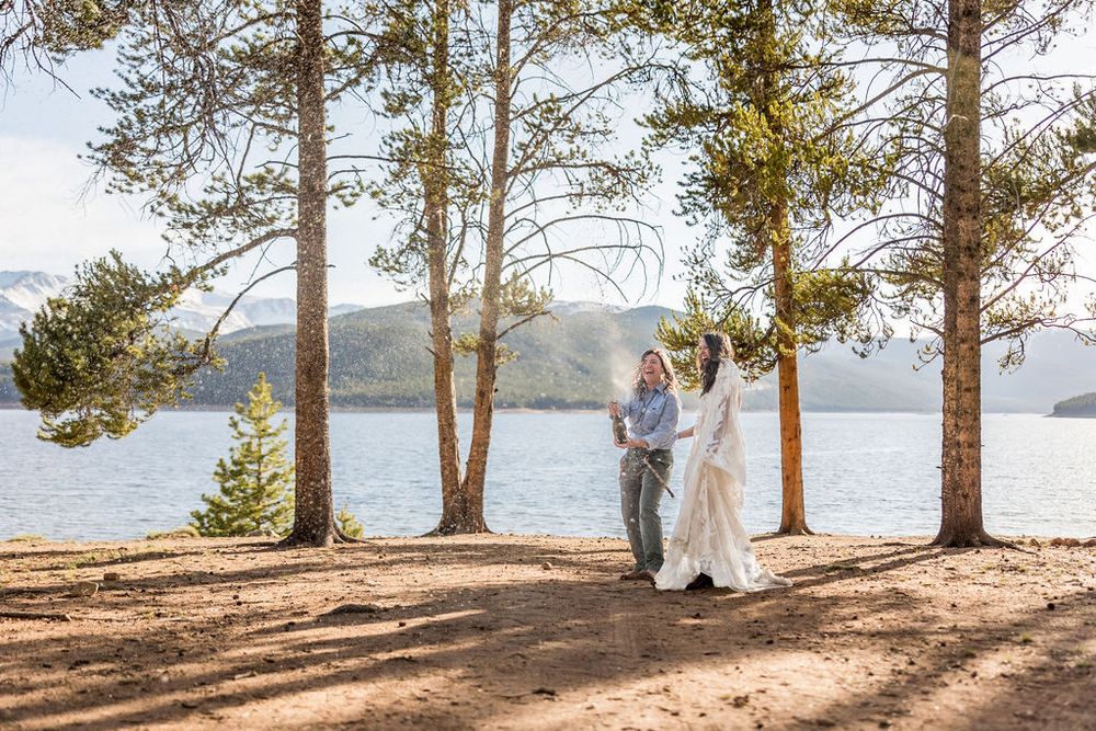 Two people standing on scenic lakeside trail among tall pine trees with mountain views in the distance.