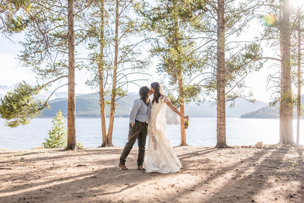 Romantic wedding couple embracing in sunlit forest by mountain lake with dramatic light rays filtering through trees.